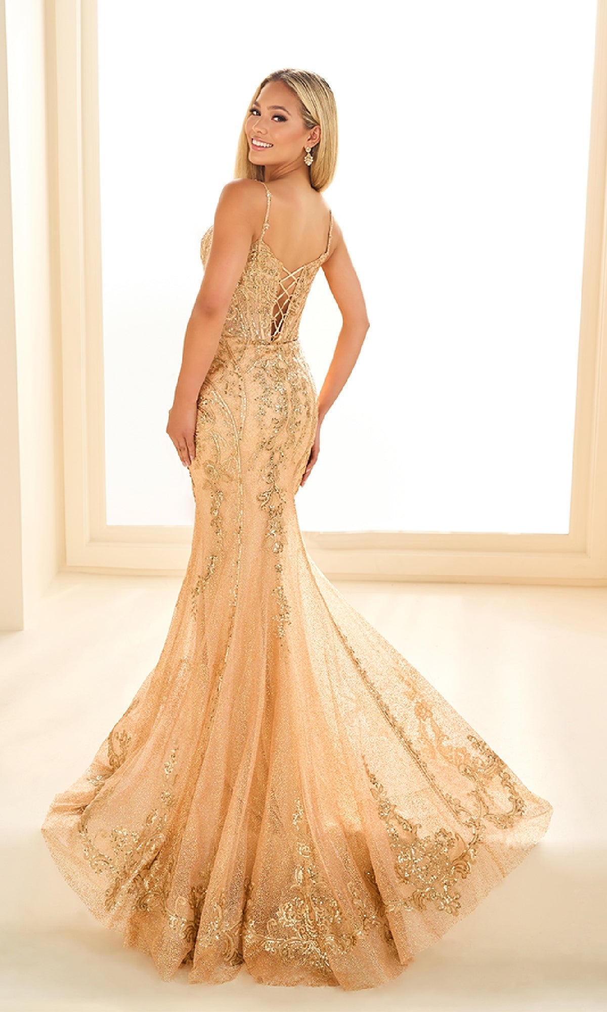 A woman smiles over her shoulder in the Ellie Wilde EW36056 gold, floor-length mermaid prom dress with lace applique details and a lace-up back.